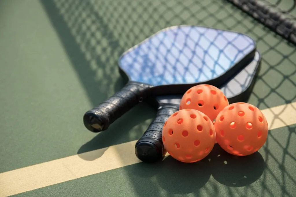 Home 1 Two pickleball paddles and three orange pickleballs sit on the court near a net, perfectly capturing the essence of #180 in an energetic match setting.