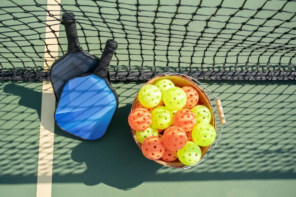 Discover the Best Pickleball Paddles for Beginners 1 Best Pickleball Paddles for Beginners: Two pickleball paddles lean against a net beside a basket filled with vibrant yellow and orange pickleball balls on court #180.