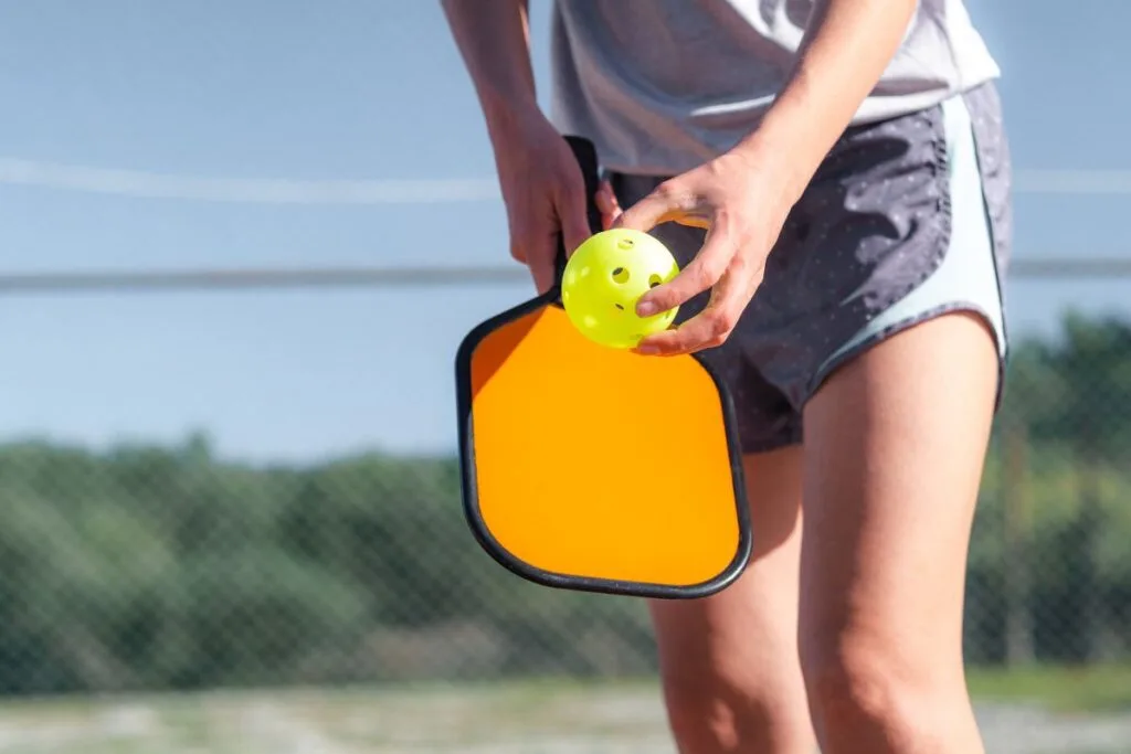 Discover the Best Pickleball Paddles for Beginners 4 A person holds a yellow pickleball and an orange paddle, one of the best pickleball paddles for beginners, as they prepare to serve on an outdoor court.
