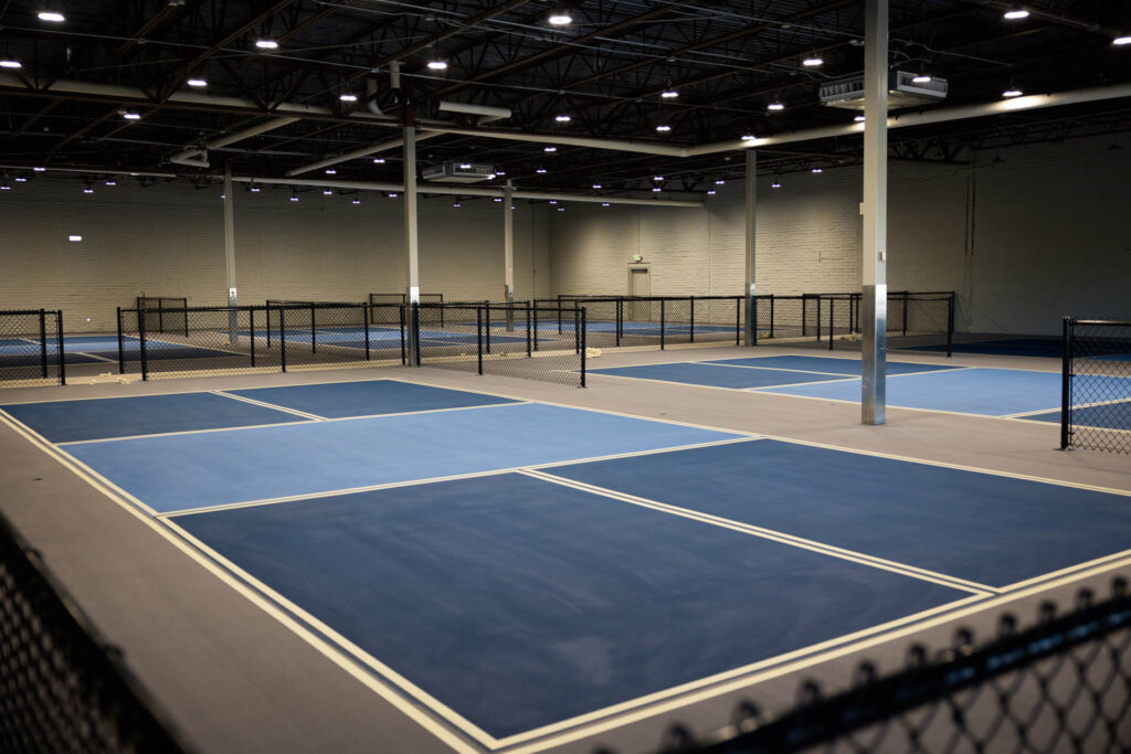 Why Pickleball Courts are the New Playground for Adults 3 Indoor facility with multiple blue pickleball courts, surrounded by black chain-link fencing, under a ceiling with exposed beams and lights. Make your reservations today and secure your spot for an event that will leave a lasting impression!