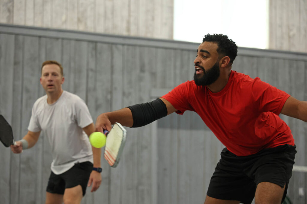Book an Event 2 Book an event at Planet Pickle: Two men playing pickleball indoors, one in a red shirt reaching for the ball with a paddle during an exciting event.