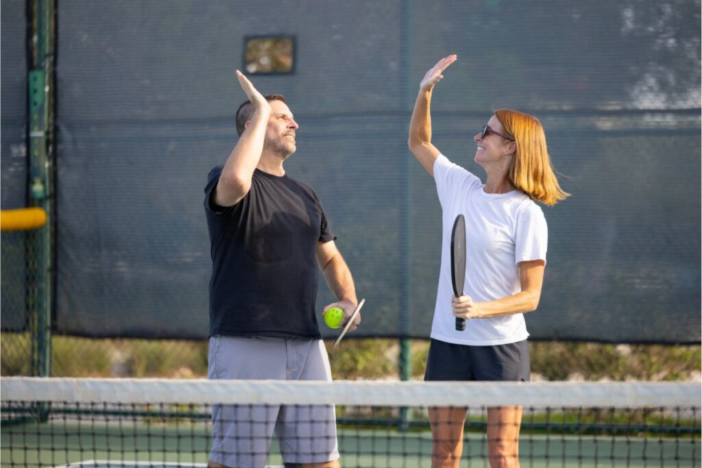 Discover the Hottest Evolving Sport: What is Pickleball 3 Two people on a pickleball court high-fiving, each holding a paddle, showcasing the camaraderie and fun inherent in this popular sport. Curious about what is pickleball? It's a dynamic game combining tennis, badminton, and ping-pong elements that's capturing hearts everywhere.