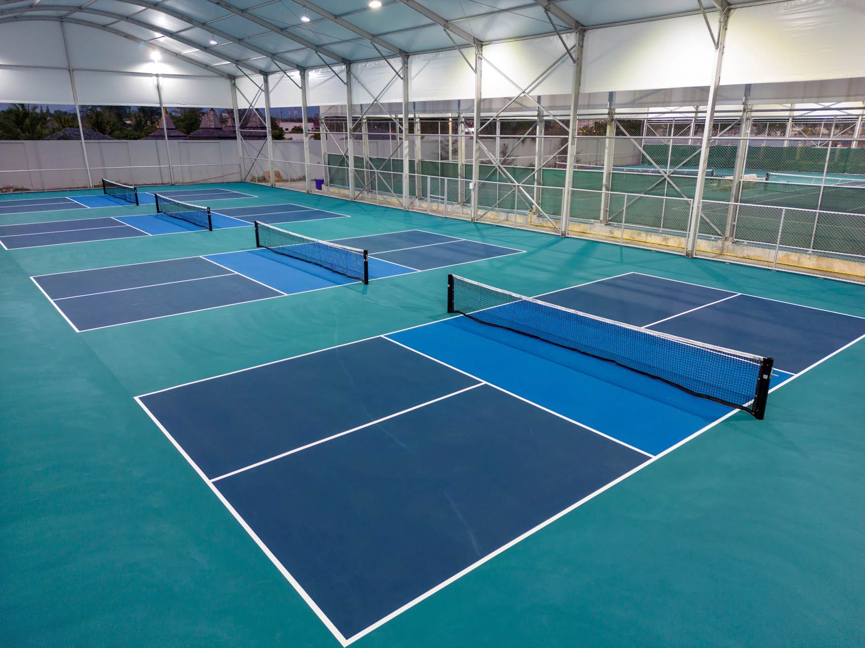 Indoor sports facility with multiple blue and green pickleball courts lined up side by side, covered by a large white roof.