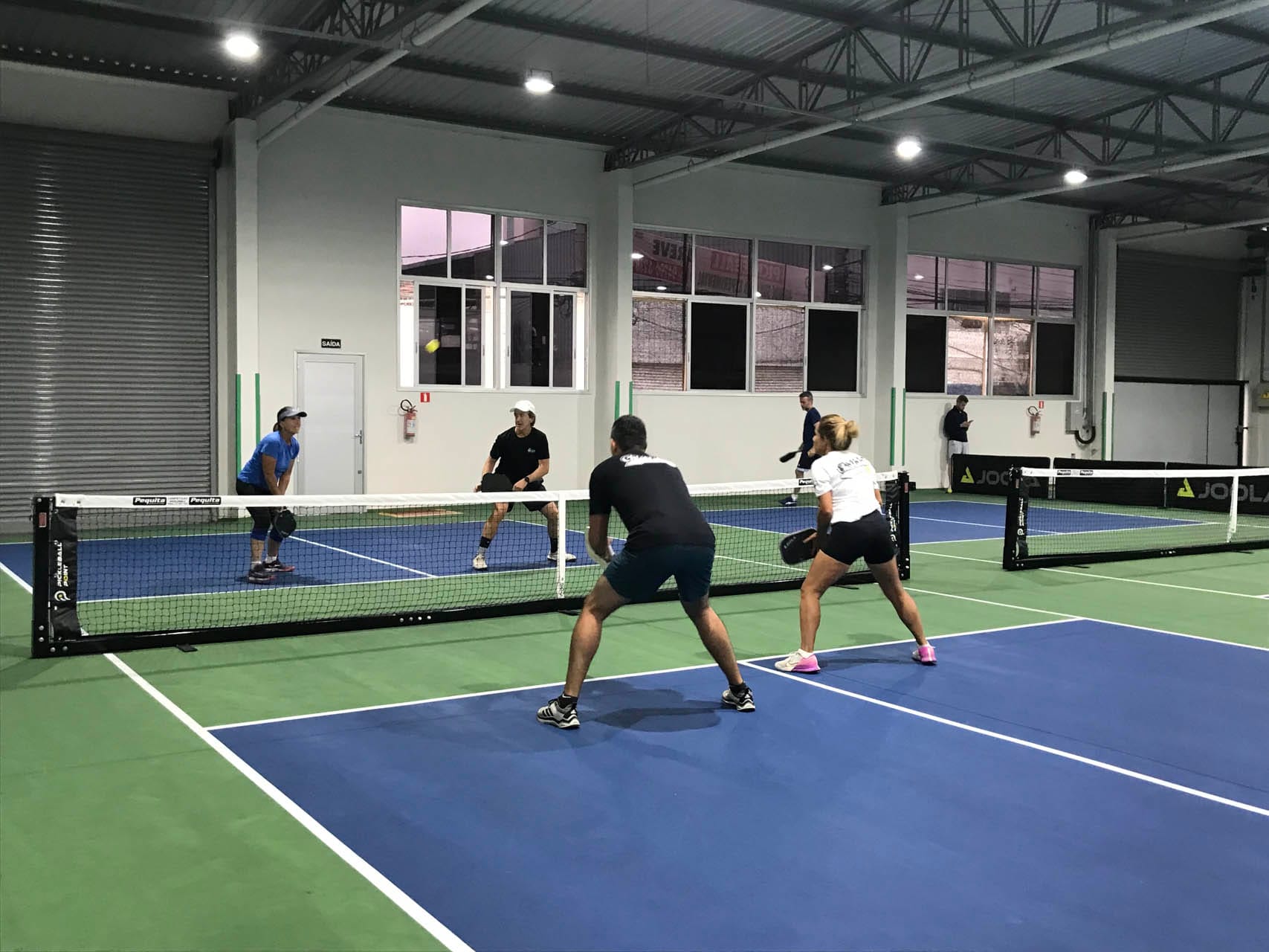 Four people playing pickleball indoors, with two players on each side of the net under artificial lighting.