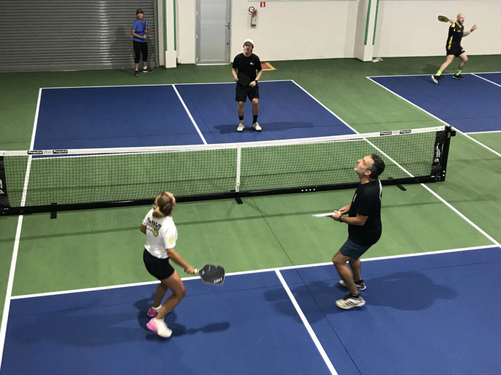 Home 2 Four people playing pickleball indoors on blue and green courts. The focus is on two players nearest the camera, preparing for a shot.
