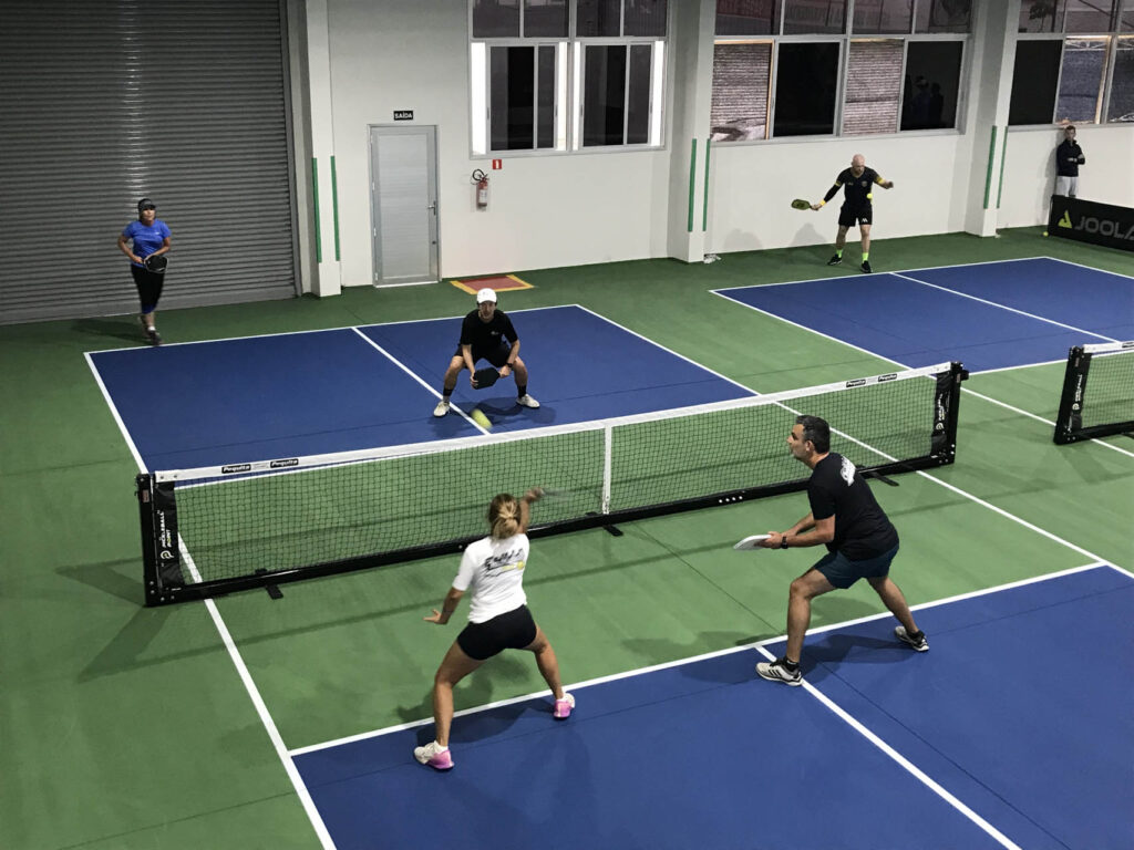 Why Pickleball Courts are the New Playground for Adults 2 Four people play pickleball on indoor pickleball courts. Two teams of two are positioned on either side of the net, each player holding a paddle.