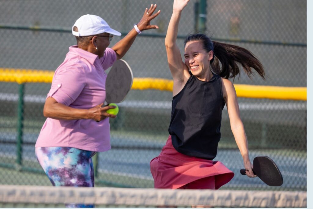 Home 8 Two women play pickleball on an outdoor court, smiling and high-fiving while holding paddles.