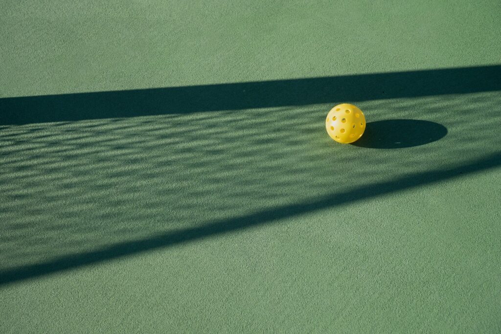 Book an Event 1 A yellow perforated ball on a green surface, partially shaded by a net's shadow.