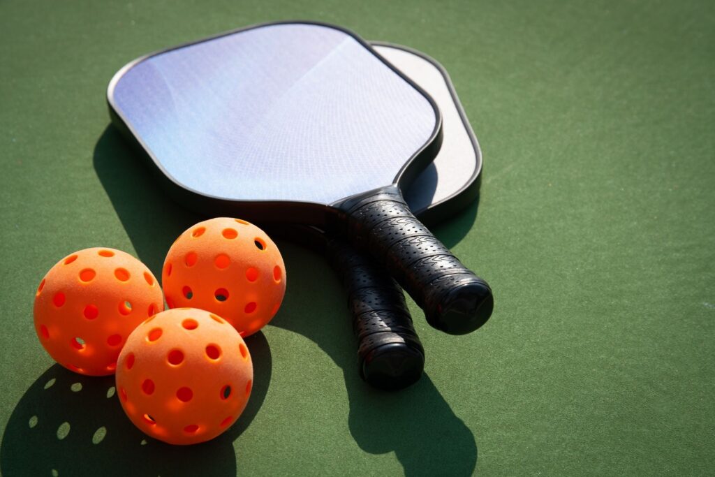 Training 3 Two pickleball paddles and three orange perforated balls on a green surface. Contact Planet Pickle in Georgia today!