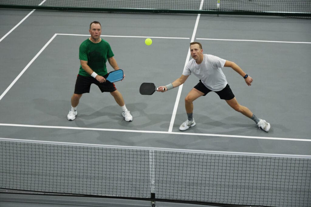 Two men engage in open play pickleball on an indoor court, both focused on the ball. One prepares to return a shot with his paddle while the other stands ready behind him.