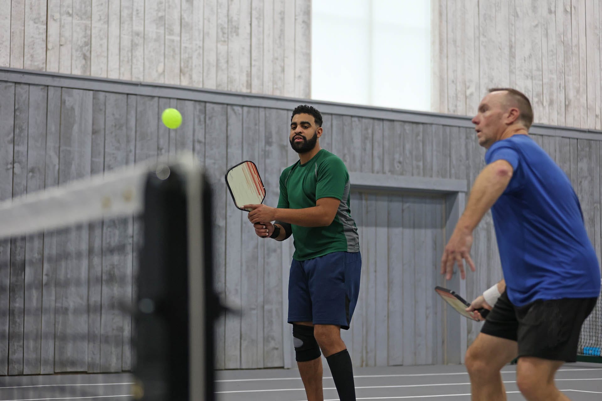 Two men engaged in open play pickleball indoors, concentrating intensely on the ball near the net. One is dressed in a green shirt and shorts, while the other sports a blue shirt and shorts.