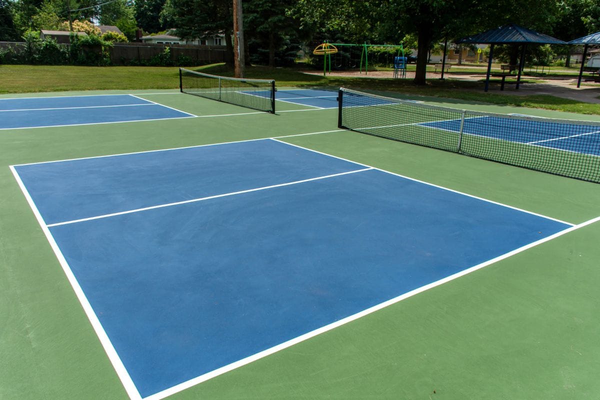 Empty outdoor pickleball courts with blue and green surfaces await players eager to explore what is pickleball, surrounded by trees and a playground in the background.