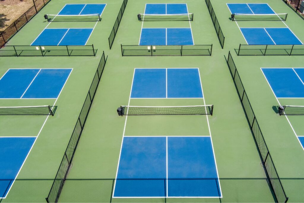 Why Pickleball Courts are the New Playground for Adults 1 Aerial view of outdoor pickleball courts with their signature blue surfaces and green borders, neatly arranged and separated by sleek black fencing.