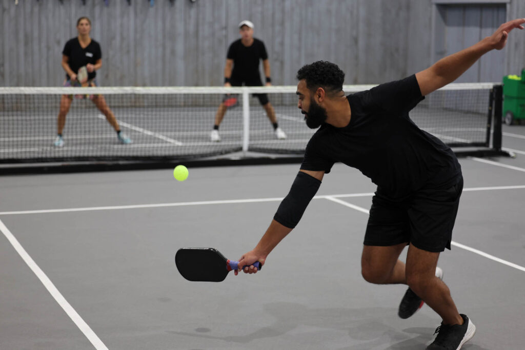 Programs 2 Three people engaging in indoor pickleball; a man in the foreground focuses intently, preparing to hit the ball during a training match, while two players wait strategically at the net on the opposite side.