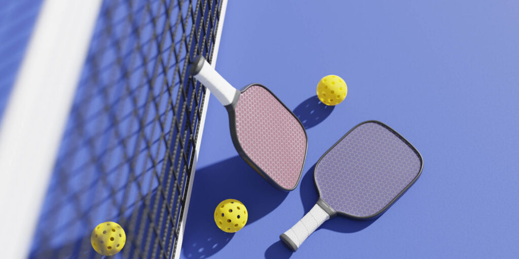 Programs 1 Two pickleball paddles and three yellow balls, essentials for any training session, lie on a blue court near the net. Whether you’re practicing for fun or preparing for an upcoming match, be sure to check the Planet Pickle calendar to plan your next session!