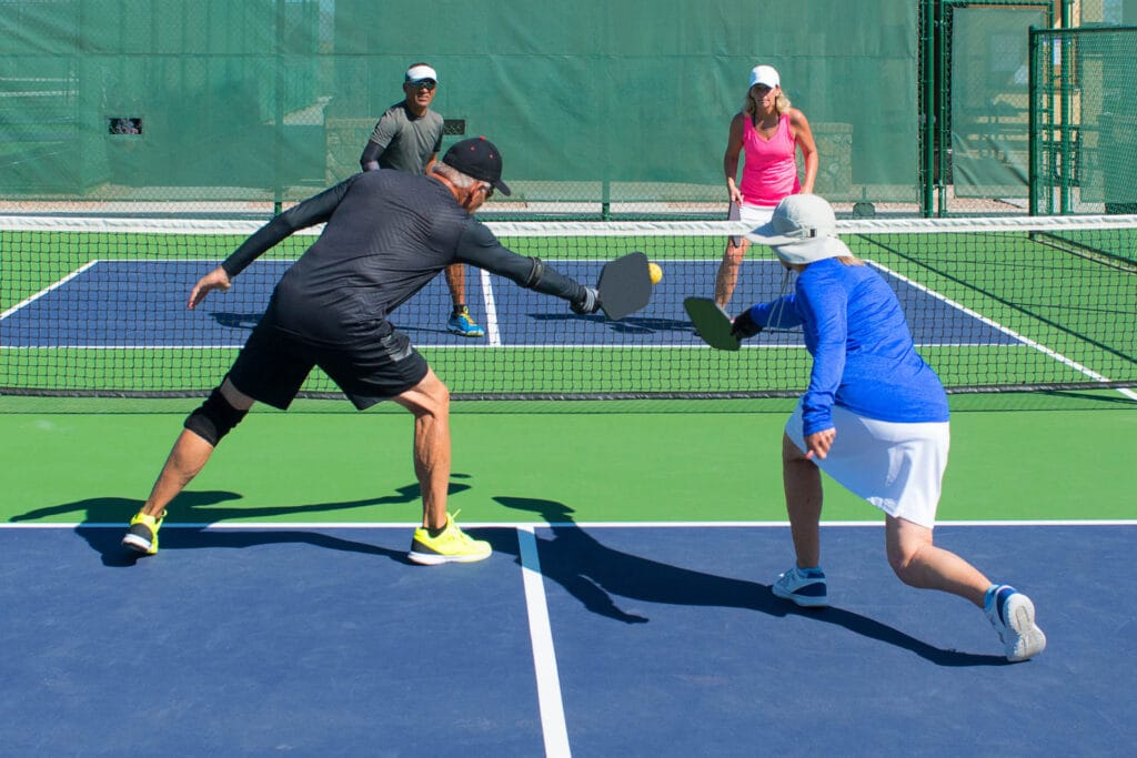 Four people play doubles pickleball on an outdoor court; two players are at the net, both reaching for the ball, while their partners watch from behind during a focused training session, perfect for private events.