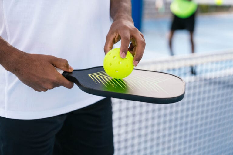 Blog 6 Best Pickleball Paddles for Beginners: Close-up of a person holding a pickleball paddle and ball on a court, with a net in the background, ready to play as reservations ensure their spot on this lively day.
