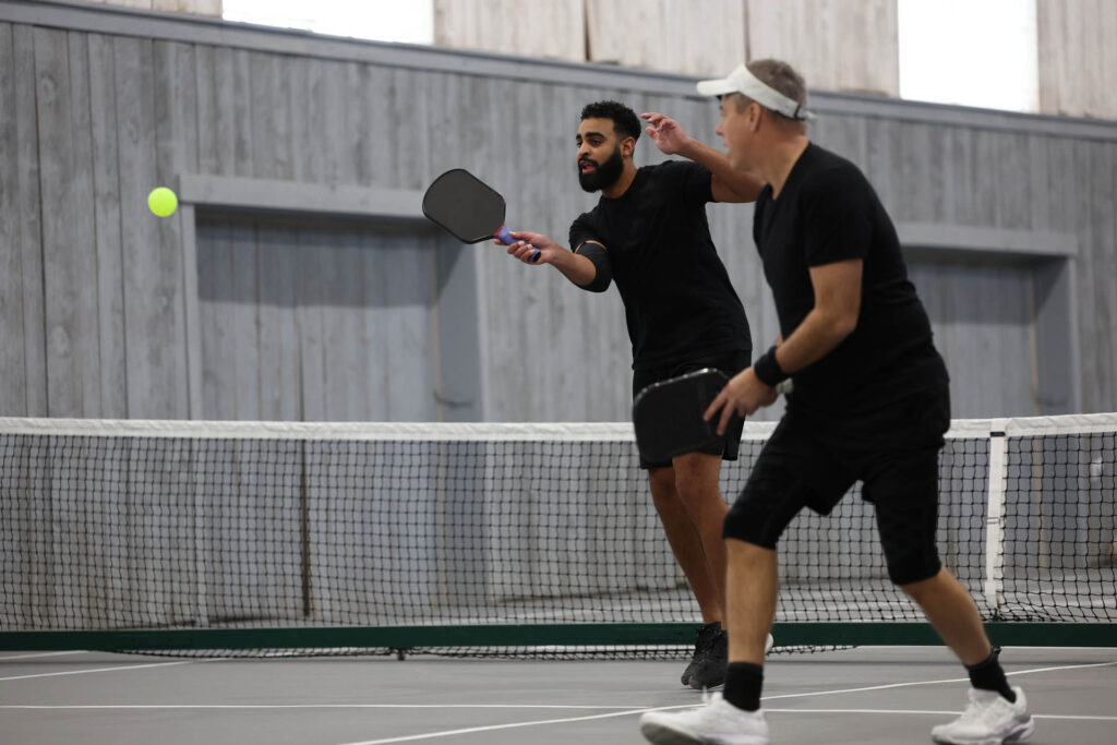 Why Pickleball Courts are the New Playground for Adults 4 Two men, amid quick footwork and swift paddles, engage in an exhilarating indoor pickleball match. Their dynamic rally is a testament to the sport's appeal, where securing pickleball courts reservations is just as crucial as perfecting a return.
