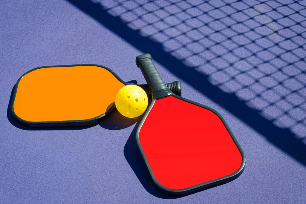Training 1 Two pickleball paddles, one orange and one red, rest on a purple court. A yellow pickleball sits between them, waiting to be served. The net casts a shadow over the surface, hinting at the excitement that follows each reservation made for this lively sport.