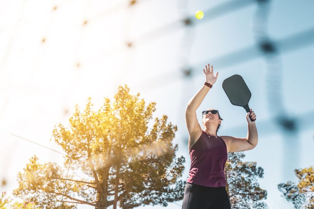 Discover the Hottest Evolving Sport: What is Pickleball 2 A person in sunglasses is ready to hit the pickleball, paddle in hand, against a backdrop of sunlit trees. For those wondering what pickleball is, it’s a dynamic mix of tennis and ping-pong played outdoors with enthusiasm and flair.
