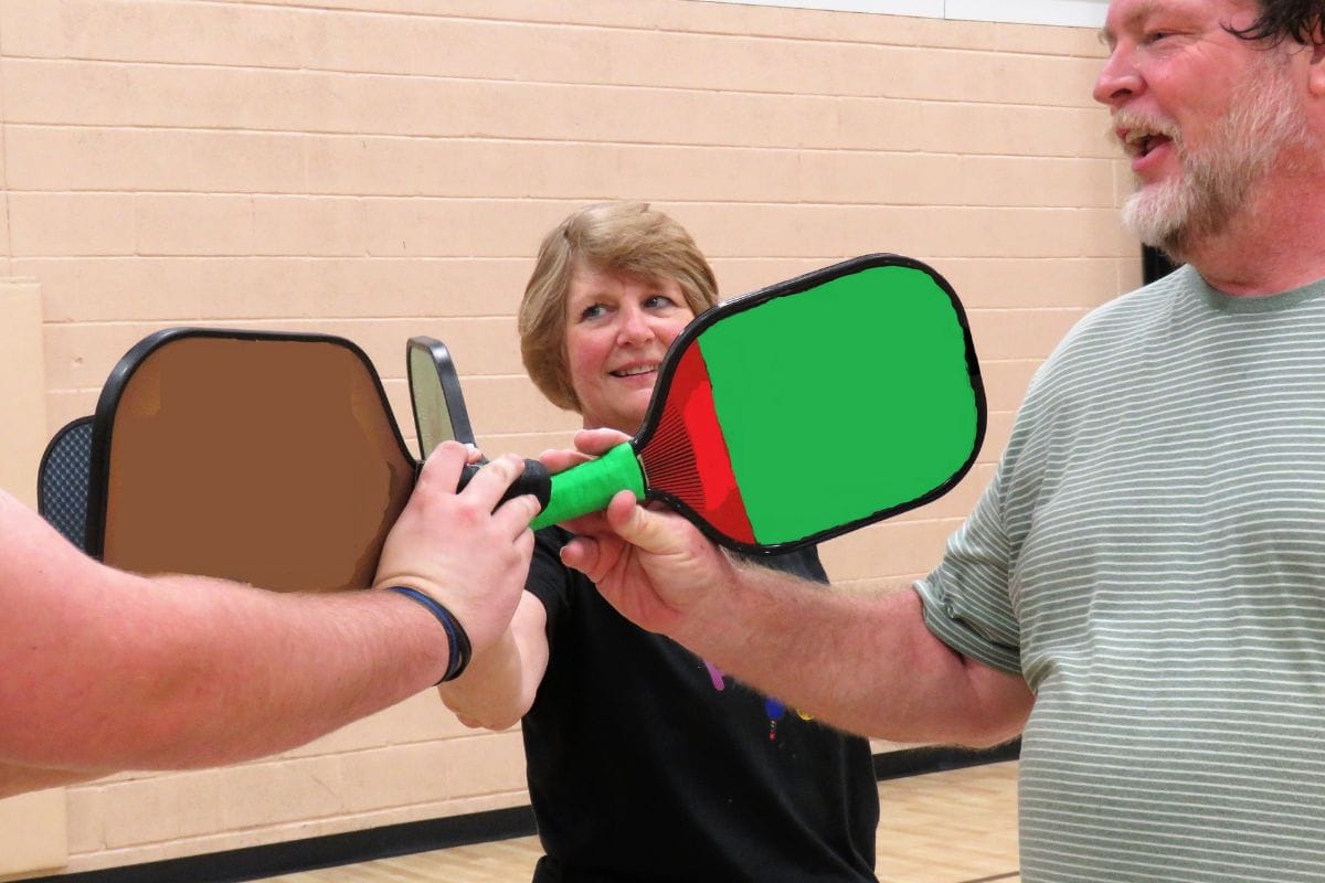 Three people stand indoors, each holding a pickleball paddle, touching them together in the center. If you're asking what is pickleball, it's this friendly gathering that showcases teamwork and joy within a game loved by many.