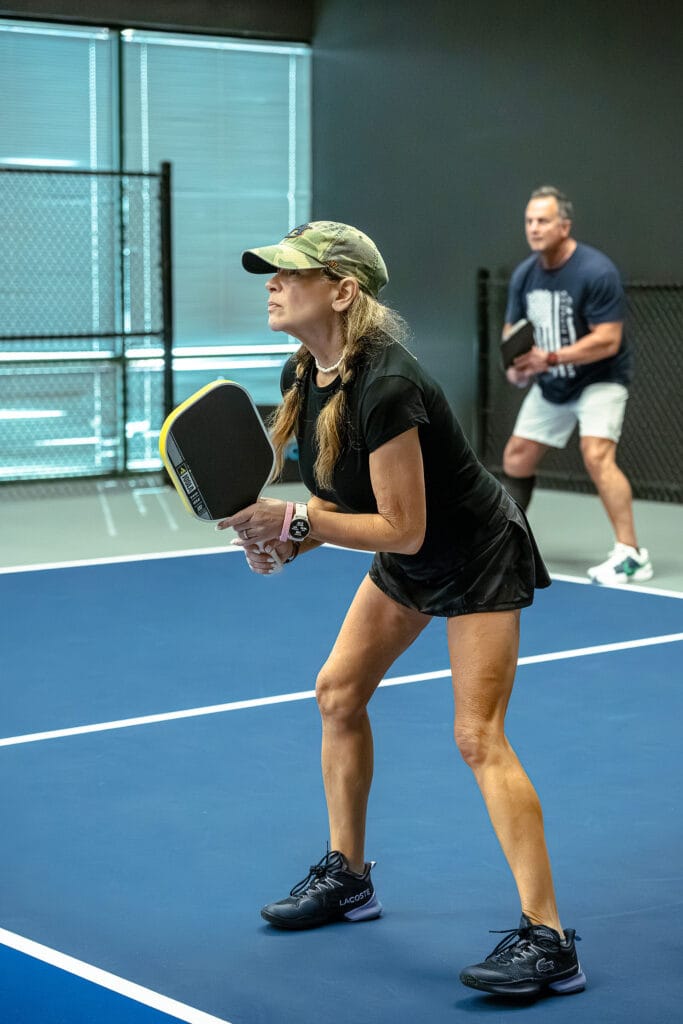 Two people stand ready with paddles on an indoor pickleball court, taking their positions along the Global Row of the court and preparing for the next play.