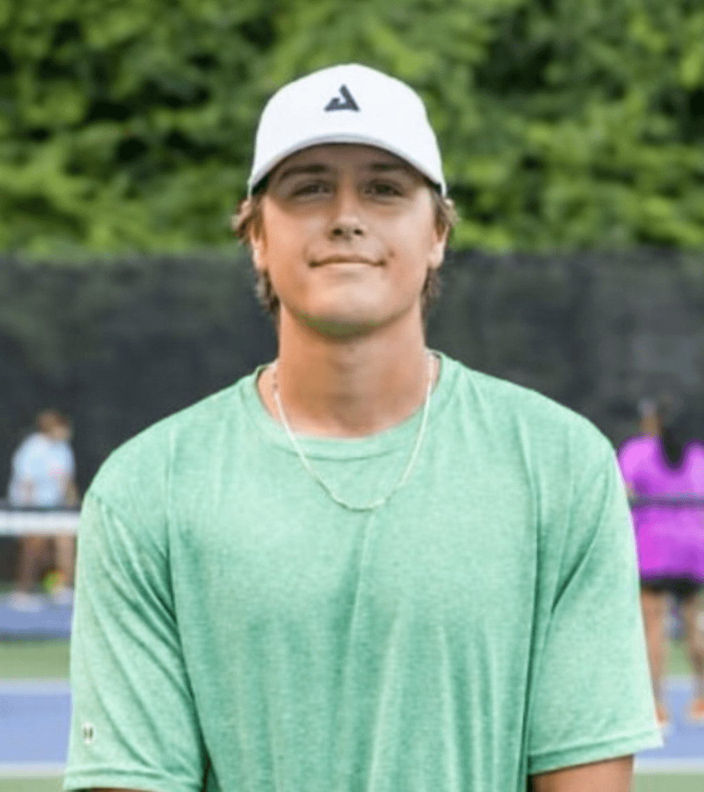 A man in a green athletic shirt and white cap stands on an outdoor tennis court, where dynamic content surrounds him with trees and other people in the background.