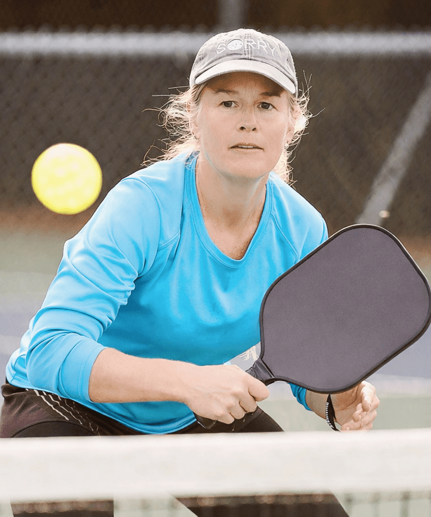 A woman wearing a blue shirt and cap plays pickleball, focusing on an incoming ball while holding a paddle near the net on an outdoor court, captured as part of a dynamic content widget showcasing active lifestyles.