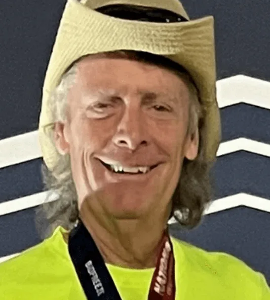 A smiling older man wearing a straw hat and neon yellow shirt with a medal around his neck stands in front of a dark background with white zigzag lines, as if he's part of a dynamic content widget display.