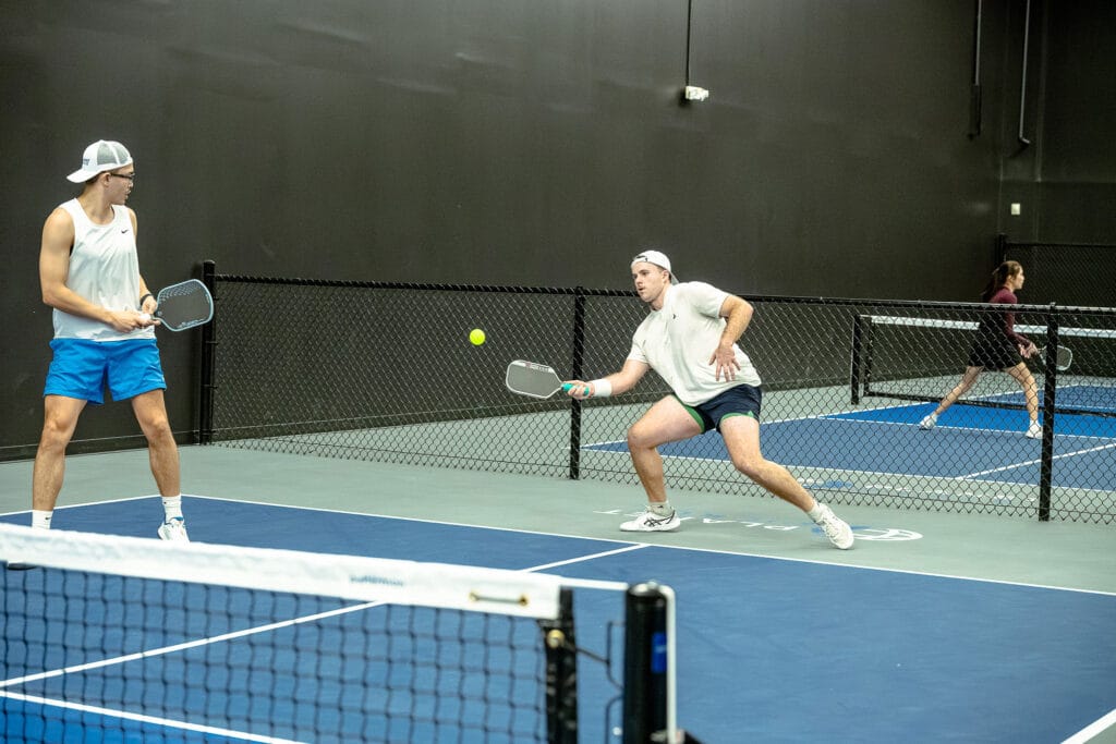 Two men play doubles pickleball indoors, preparing to hit a ball mid-air; in the background, another player is visible on a separate court near a dynamic content widget.