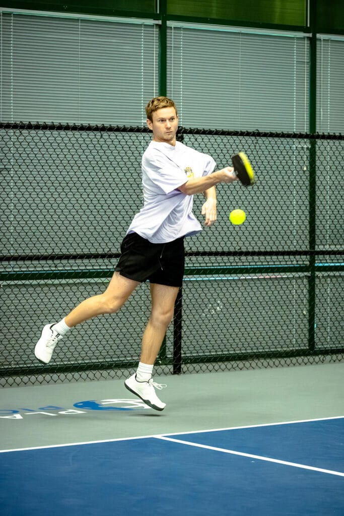 A man in athletic wear jumps to hit a yellow pickleball with a paddle on an indoor court, while a dynamic content widget displays game stats beside the fence and windows in the background.