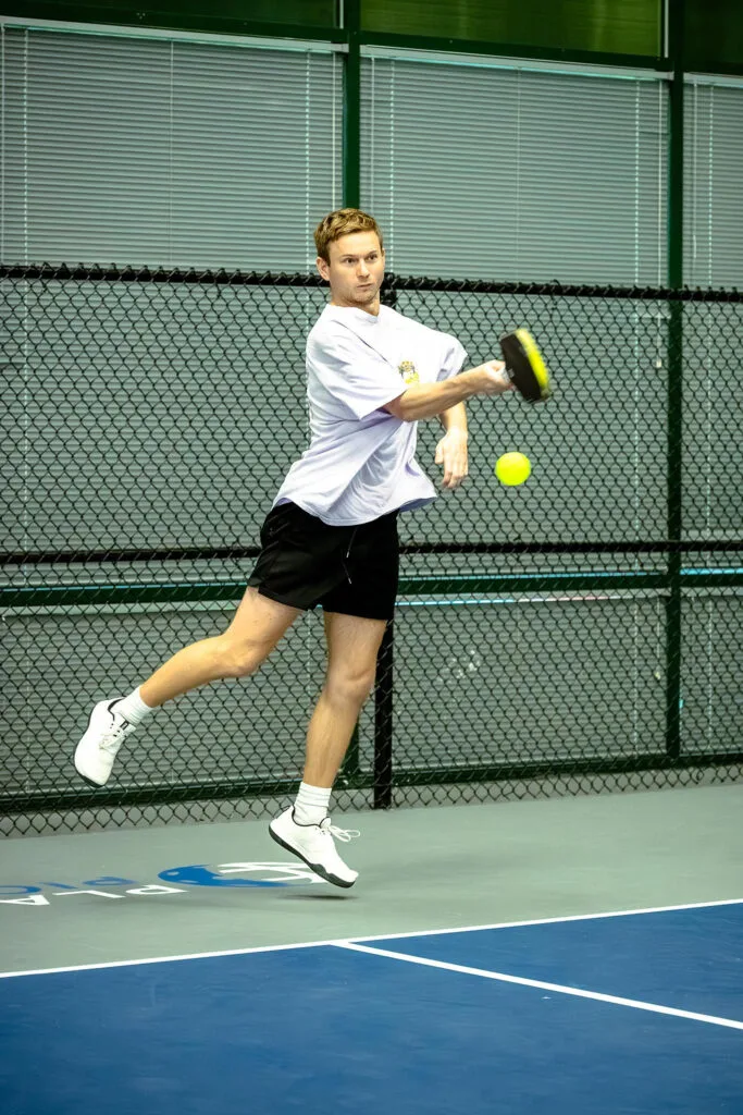 A man in athletic wear jumps to hit a yellow pickleball with a paddle on an indoor court, while a dynamic content widget displays game stats beside the fence and windows in the background.
