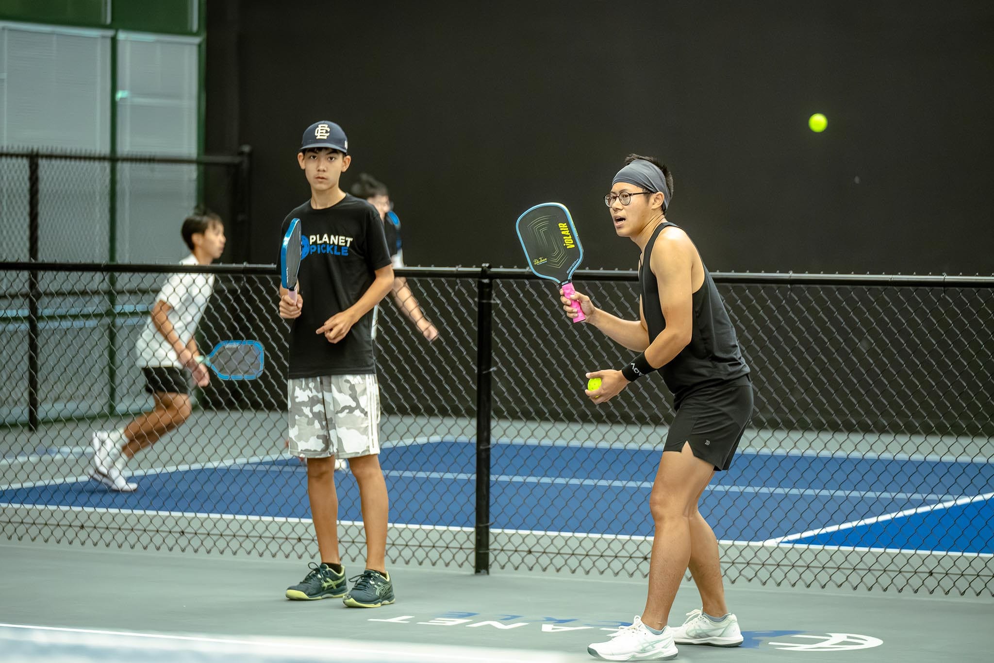 Two pairs of people play pickleball on indoor courts; the pair in the foreground, equipped with pickleball paddles, prepares to serve, while the pair in the background is caught mid-rally.