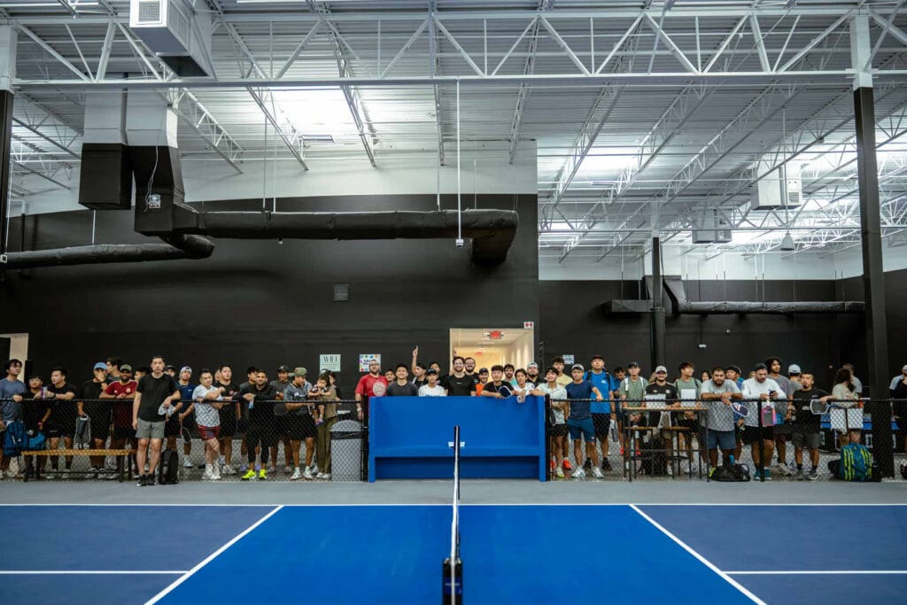 A group of people stands behind a barrier, watching an indoor pickleball court&mdash;often called a playground for adults&mdash;under a high ceiling with exposed beams and ducts.
