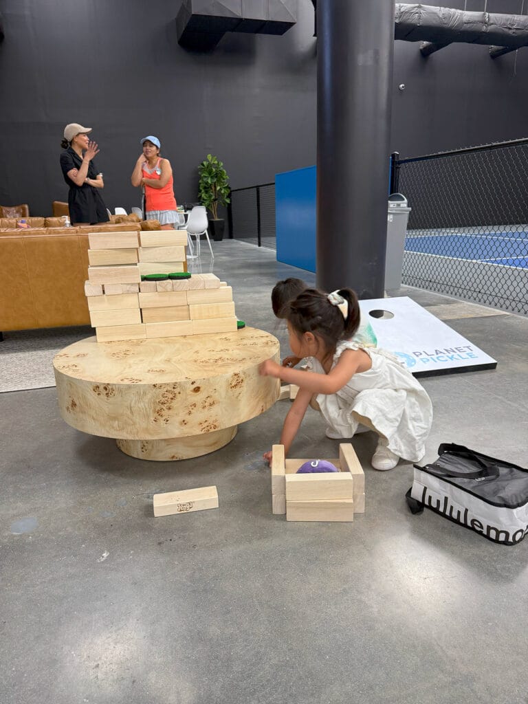 Home 27 Two young children play with large wooden blocks on a round wooden table while two adults talk in the background inside a spacious indoor area, creating an inviting scene perfect for a homepage or newsletter feature.
