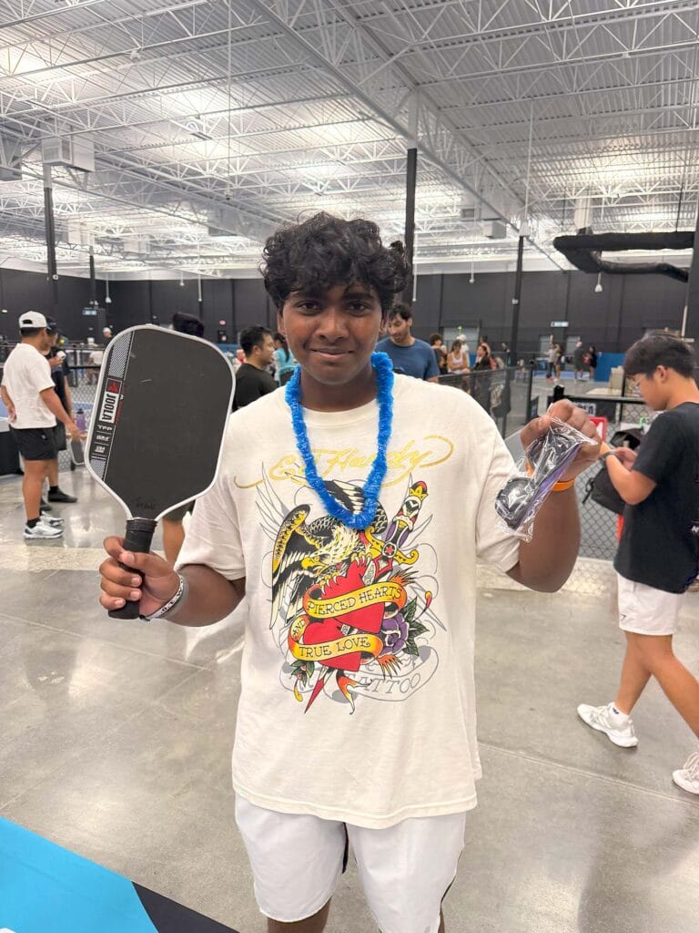 Home 29 A person holding a pickleball paddle and a prize, wearing a graphic t-shirt and a blue lei, stands on an indoor court with other players in the background—perfect for your homepage or next newsletter feature.