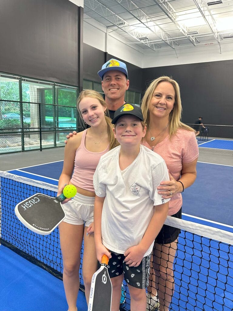Home 25 A family of four stands on an indoor pickleball court, holding paddles and smiling at the camera—perfect for your newsletter or homepage.