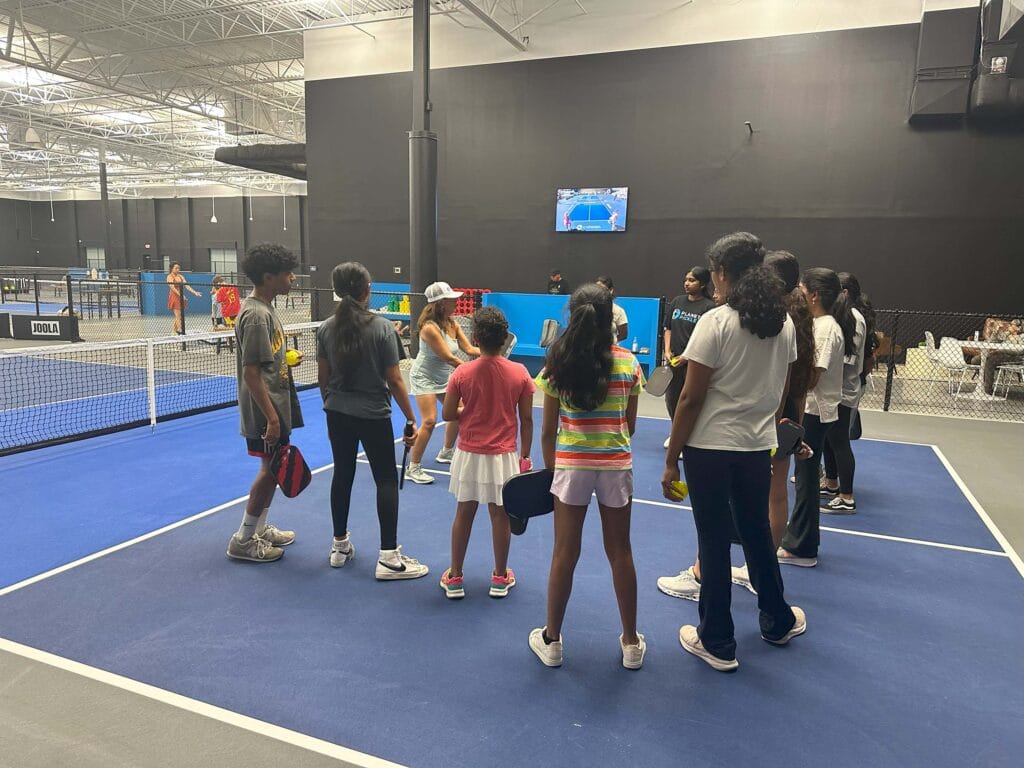 Home 34 A group of children and teens stand in a circle on an indoor pickleball court, holding paddles and facing an instructor—perfect for your newsletter or homepage feature.