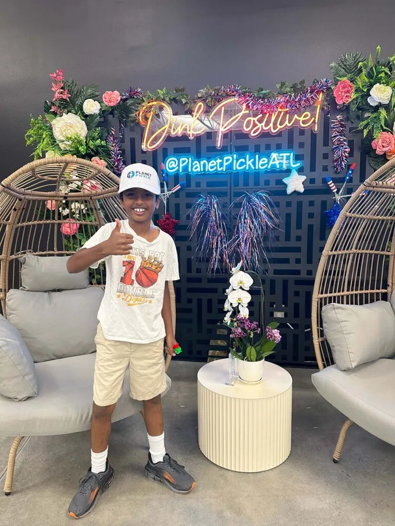 Home 9 A boy in a white t-shirt and hat stands smiling with a thumbs up in front of a decorative floral wall with neon signs reading “Dink Positive” and “@PlanetPickleATL,” perfect for the Homepage or Newsletter.