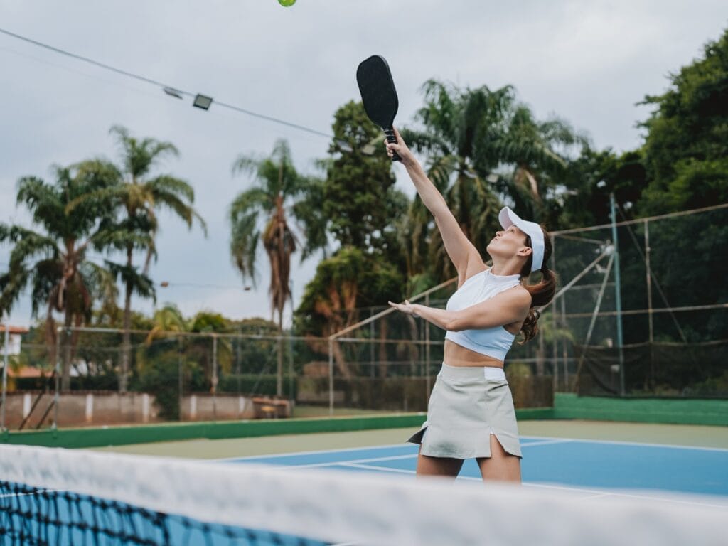 Basic Pickleball Rules for New Players 3 A woman playing pickleball prepares to hit an overhead shot on an outdoor court surrounded by palm trees, showcasing her skills while following basic pickleball rules.