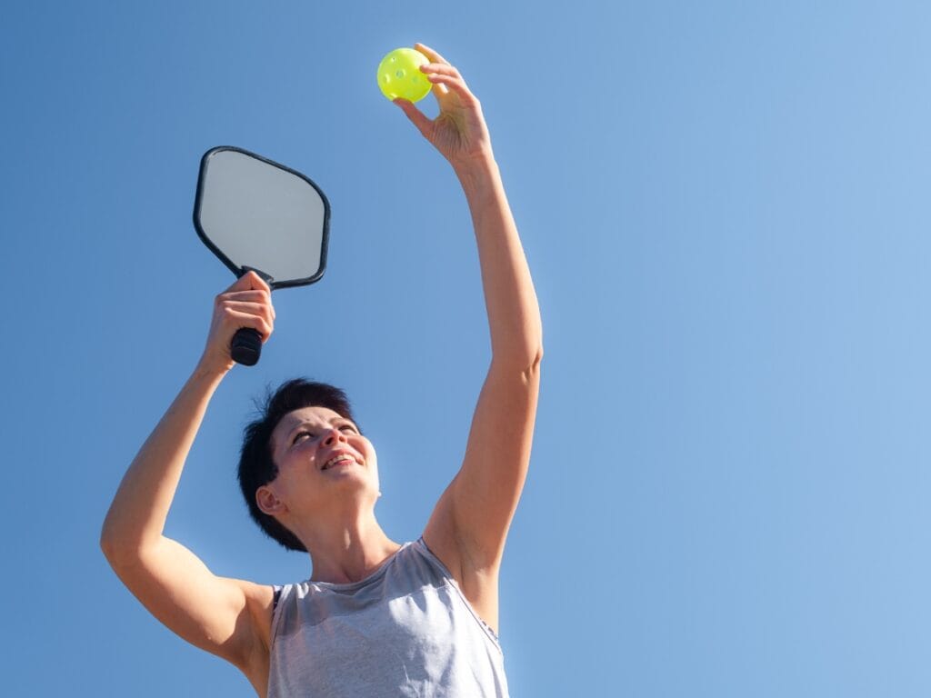 Basic Pickleball Rules for New Players 5 A person prepares to serve a yellow pickleball with a paddle, holding both arms raised against a clear blue sky, demonstrating Basic Pickleball Rules before the game begins.