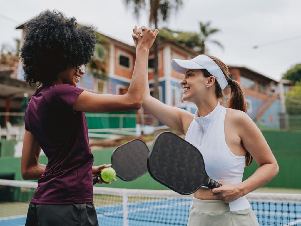 Basic Pickleball Rules for New Players 7 Two women stand on a tennis court, high-fiving with pickleball paddles in hand—one also holding a ball—celebrating after following basic pickleball rules.