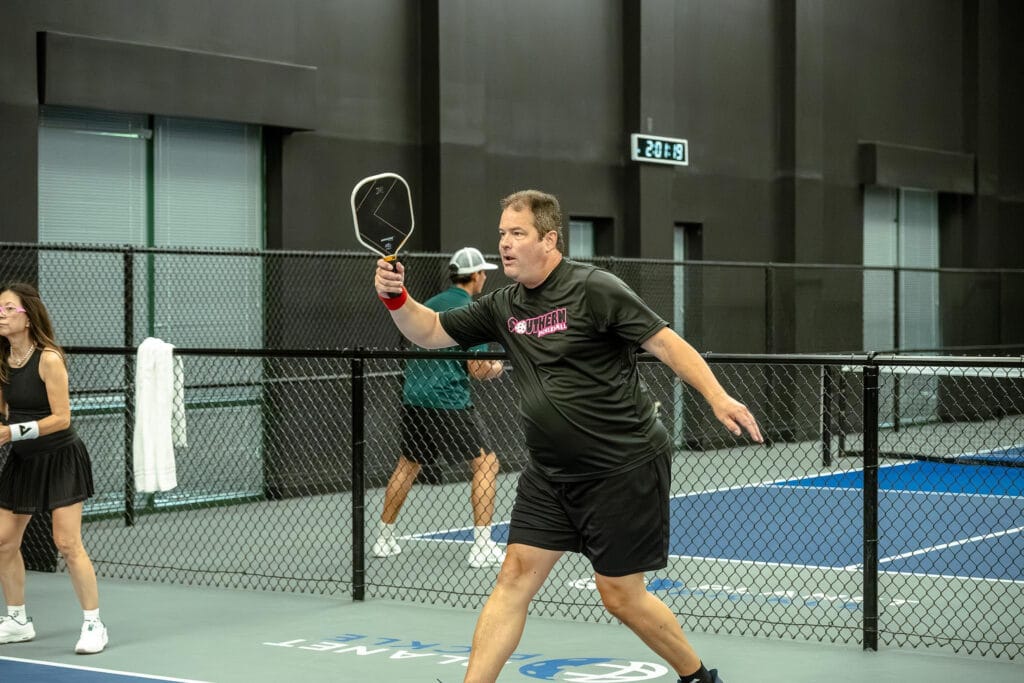 How to Play Pickleball: A Beginner’s Guide 6 A man wearing a black shirt prepares to hit a pickleball on an indoor court, illustrating how to play in this beginner’s guide, with other players visible in the background.