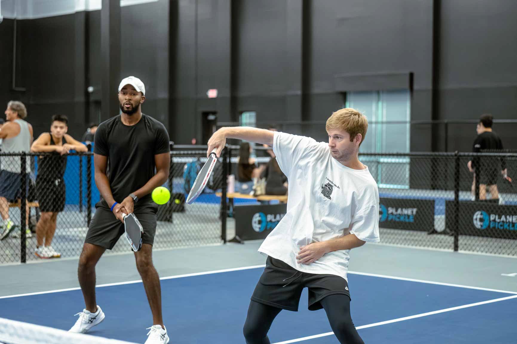 Two men play basic pickleball doubles indoors, with one hitting the ball while the other stands ready; other players&mdash;possibly new players learning Pickleball rules&mdash;watch in the background.