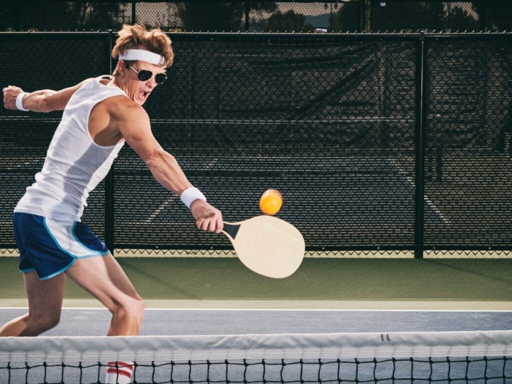 A man wearing sports attire and a headband plays pickleball, swinging a paddle to hit a ball over the net on an outdoor court, capturing why pickleball is so popular among active enthusiasts.