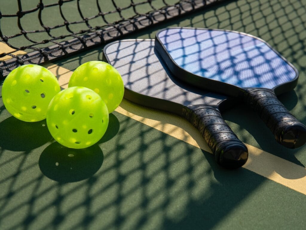 Two pickleball paddles and three bright yellow pickleballs rest on an outdoor court near a net, with shadows cast on the surface—capturing why pickleball is so popular among players of all ages.