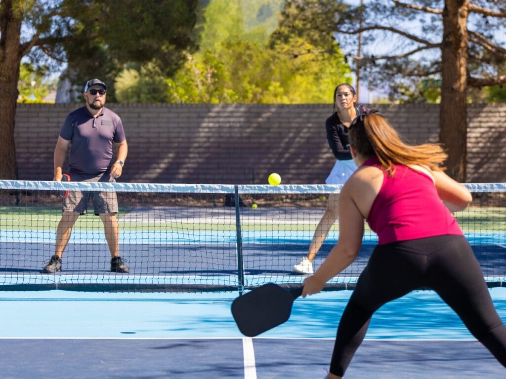 Three people play pickleball on an outdoor court, with two on one side of the net and one on the other, showing why pickleball is so popular as the yellow ball approaches the net in this energetic match.