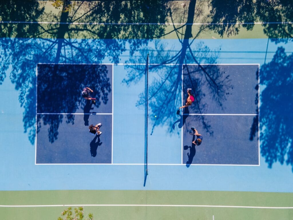 Aerial view of four people playing doubles tennis on a blue court with tree shadows visible—capturing the social energy often seen in today’s racquet sports and hinting at why pickleball is so popular.