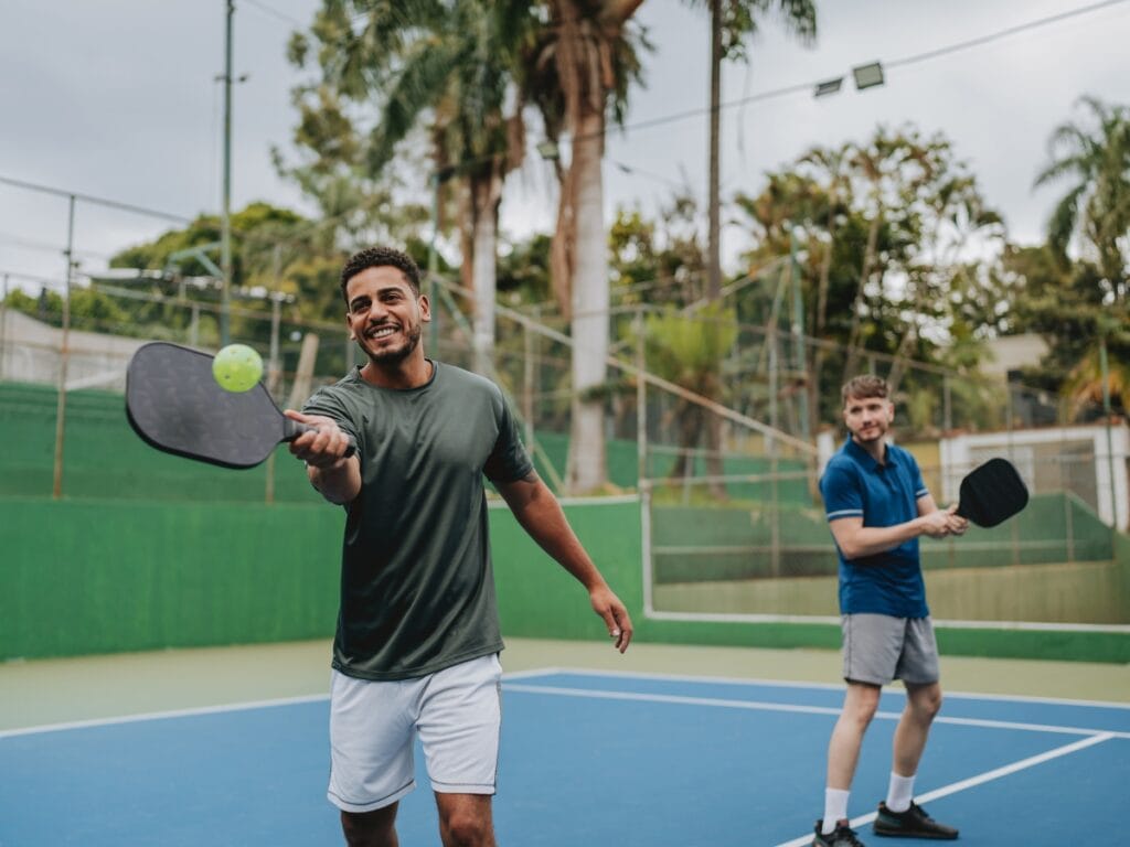 Two men playing pickleball on an outdoor court highlight the fast-paced action typical of pickleball vs tennis; one player is poised to hit the ball with his paddle as the other gets ready in the background.