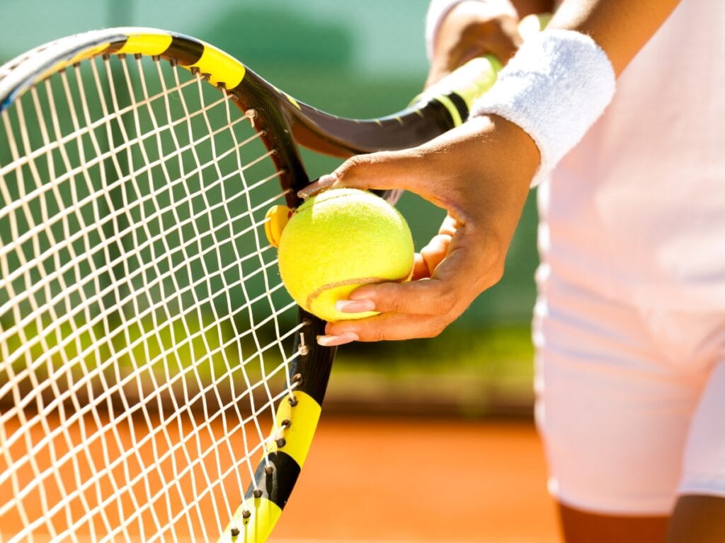 A person in white sportswear prepares to serve by placing a tennis ball against the strings of a tennis racket on an outdoor court, highlighting the classic serve technique seen in pickleball vs tennis matches.
